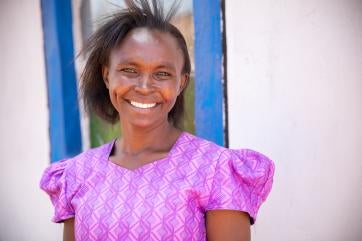 Woman smiling outdoors in front of a white wall with blue window frame.