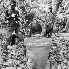 A person stands in a wooded area using a mobile device while another person sits on the ground and a third person stands nearby smiling.