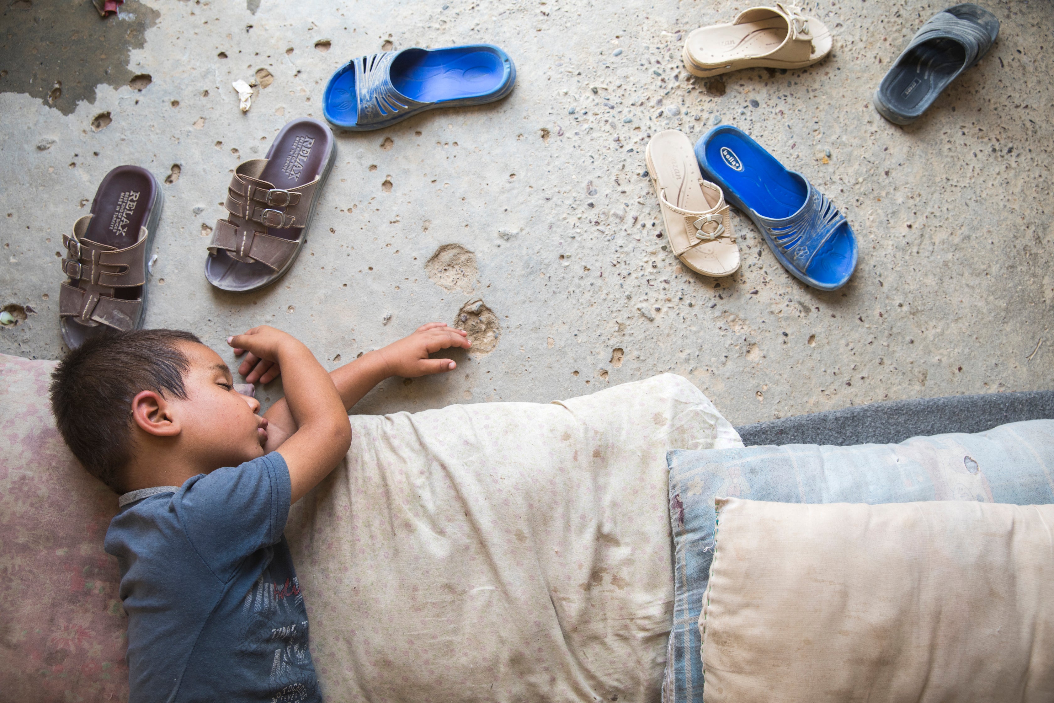 Iraqi child naps on the dirt floor inside tent, dusty sandals splayed beside him.