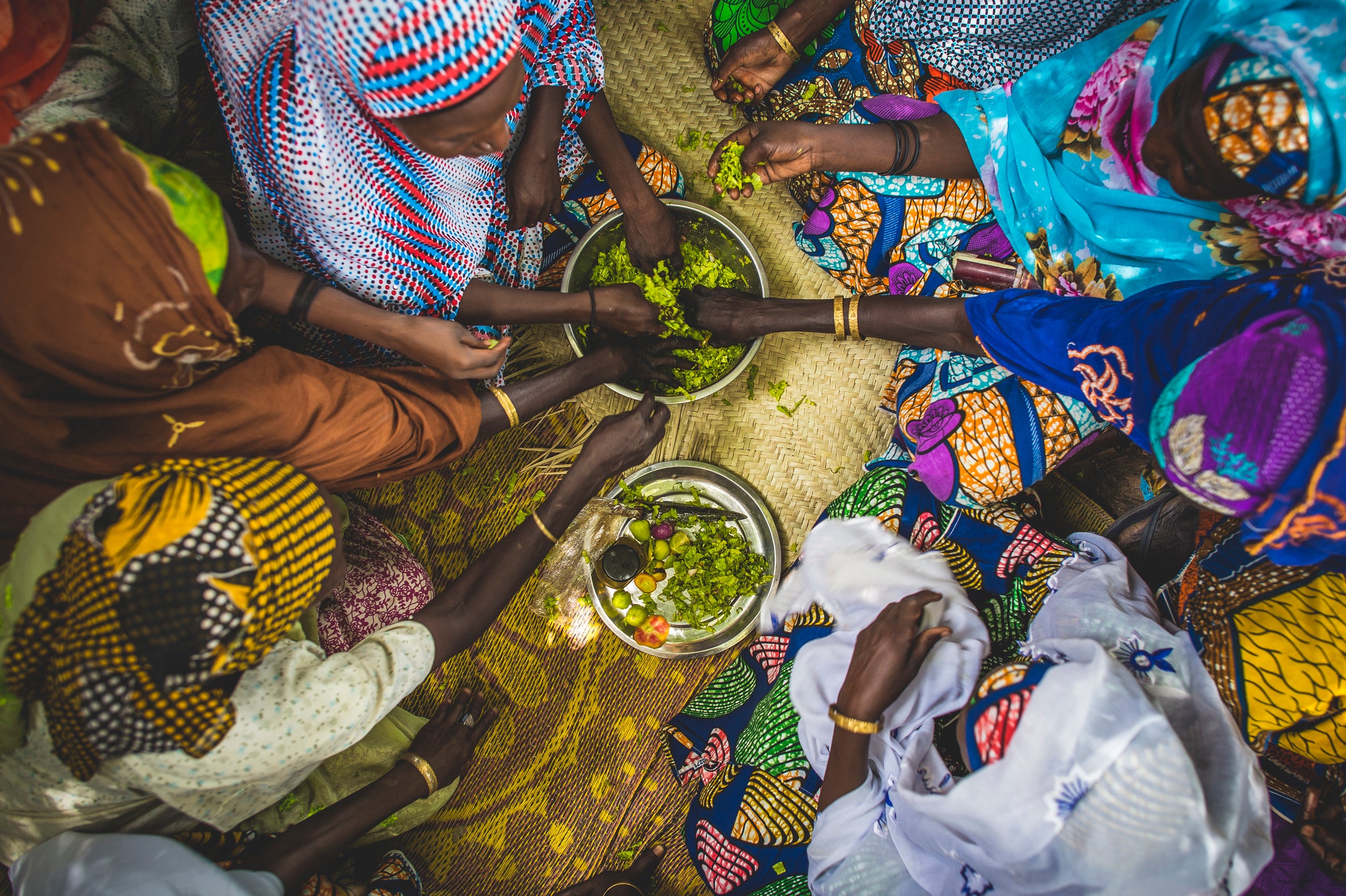 Women in colorful clothing reaching in to share food from a bowl at center in Niger. Photo: Sean Sheridan for Prosper Global