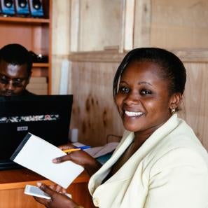 Lucy at a counter across from a man on a computer, smiling and holding a cell phone, notepad and pen.