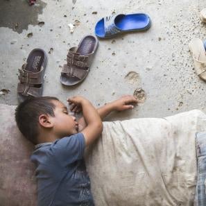 Iraqi child naps on the dirt floor inside tent, dusty sandals splayed beside him.