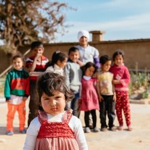A group of children in lebanon smiles and snickers in the background as one young girl approaches the camera inquisitively 