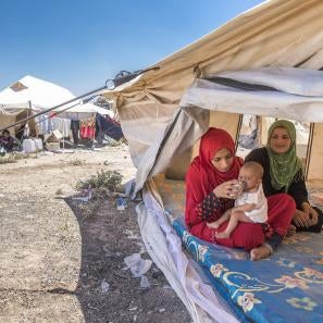 Two women sit beneath a white canvas tent. one is holding and feeding a baby on her lap.