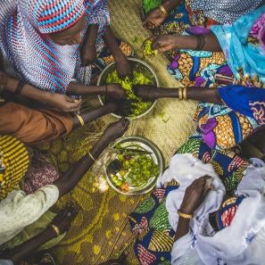 Women in colorful clothing reaching in to share food from a bowl at center in niger. photo: sean sheridan for prosper global