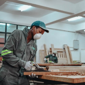 A person uses wood working tools while wearing a face mask in a room with other face masked workers.